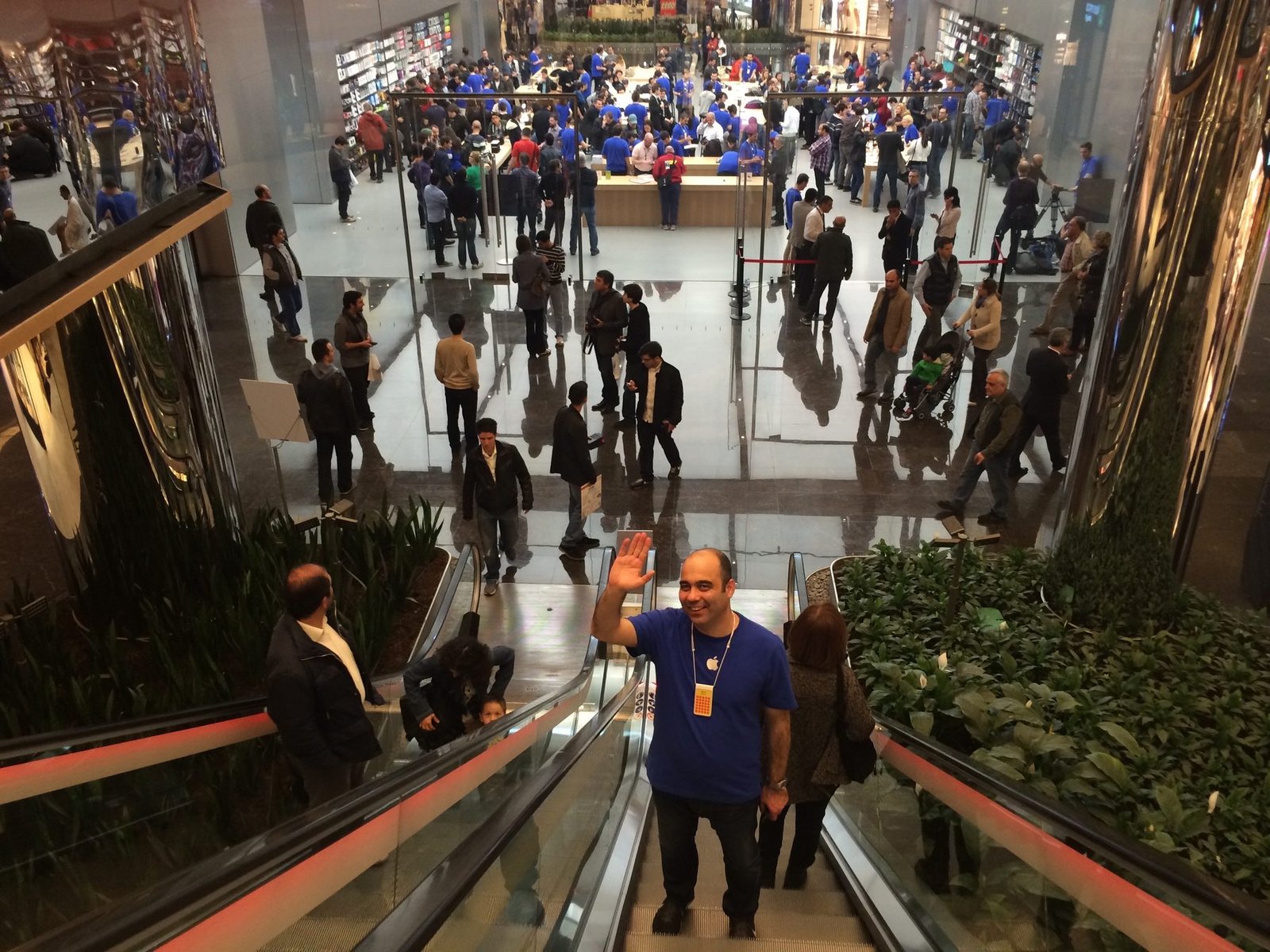 Baha Cinar waving from the escalator inside Apple Zorlu Center on opening day, the store floor crowded with customers and blue-shirted staff behind him